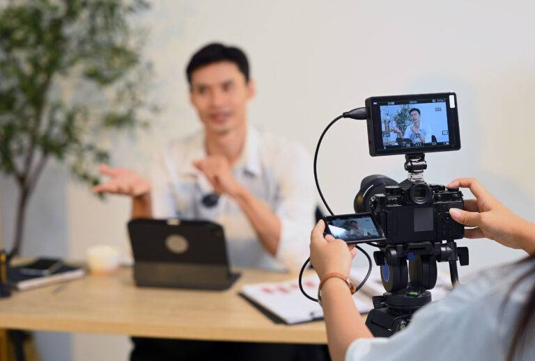 A young man sits at his desk being filmed by a colleague on a DSLR camera