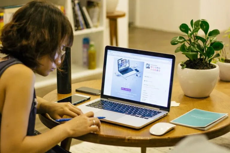 A young woman seated is working on her laptop, there is a book and a dark green potted plant on the desk