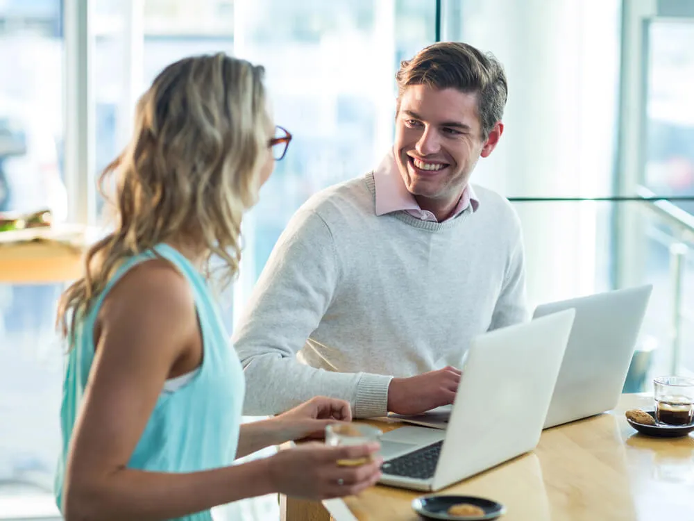 On site training - a male trainer sits next to a female student in a modern office, there is a large window behind them, they both have laptop computers open and are happily in conversation